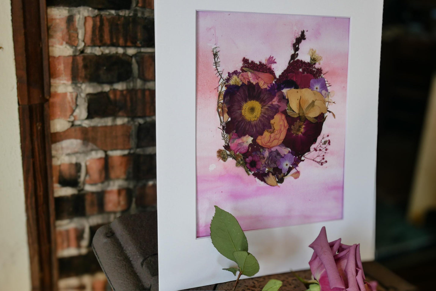 a framed artwork of a heart-shaped arrangement of dried flowers, including purple and yellow blooms, set against a pink background.