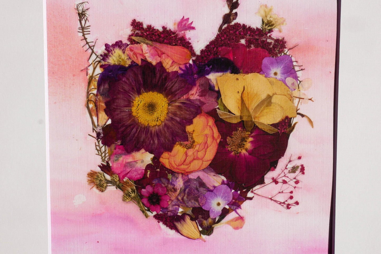 A heart-shaped arrangement of various colorful flowers, including purple, yellow, and pink blooms, against a pink background.