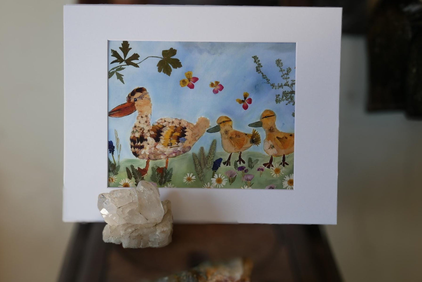 a vibrant painting of a duck family in a grassy field, with two adult ducks and two ducklings. The painting is set against a backdrop of flowers and leaves, creating a lively and colorful scene.