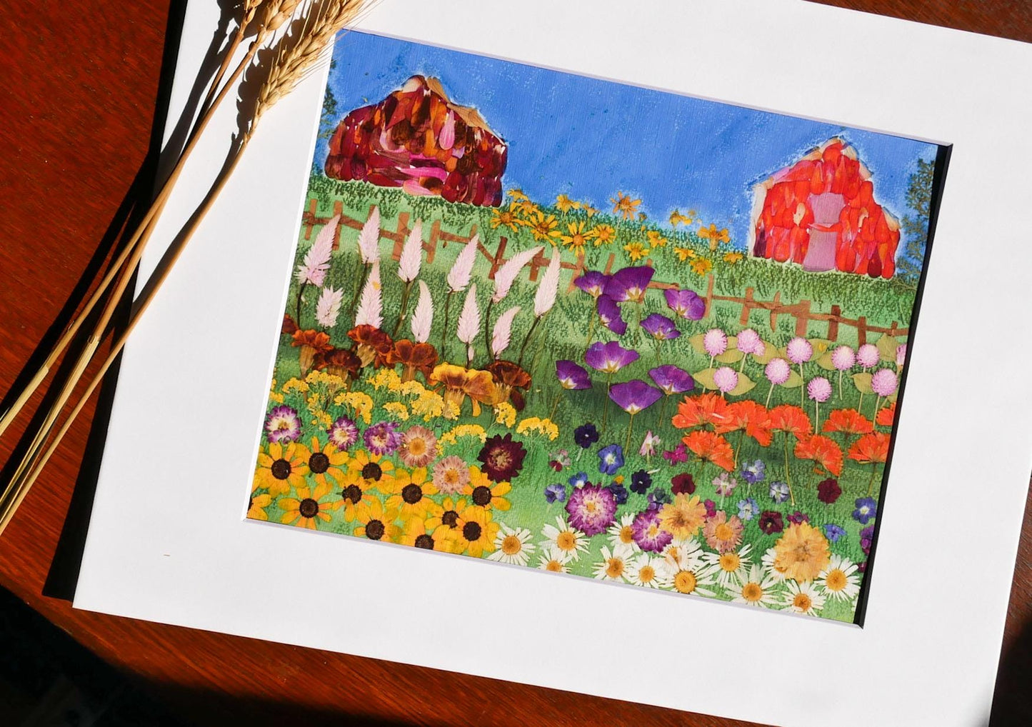 The image is a vibrant painting depicting a lush garden filled with a variety of colorful flowers, including sunflowers, daisies, and other blooms. The painting is framed and placed on a wooden surface, with a few wheat stalks visible in the background.