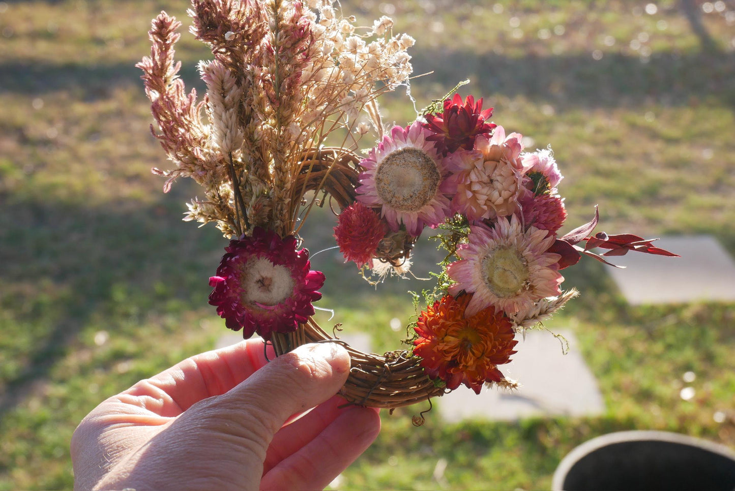 A hand holding a wreath made of dried flowers and grasses against a blurred background of a grassy field.