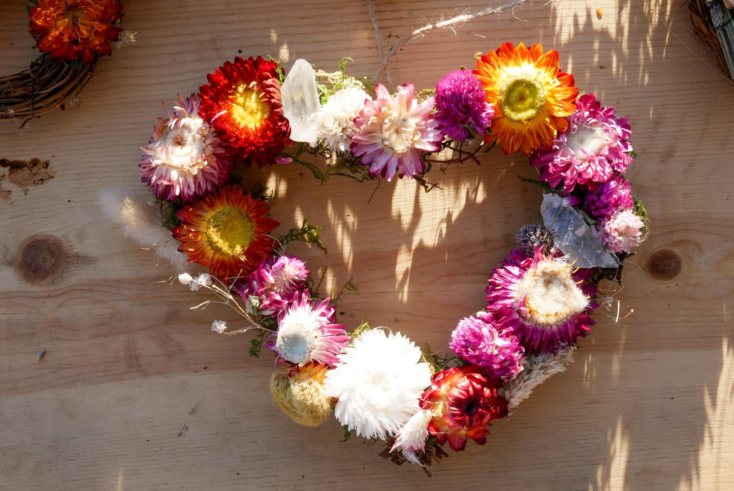 A heart-shaped wreath made of various colorful flowers, including red, orange, pink, white, and purple, is displayed on a wooden surface.