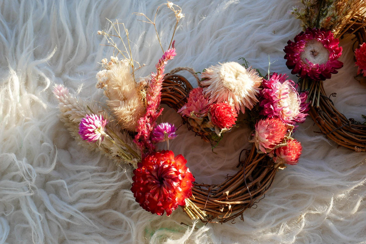 A wreath made of dried flowers and branches, with a mix of pink, red, and white flowers, arranged on a white furry surface.
