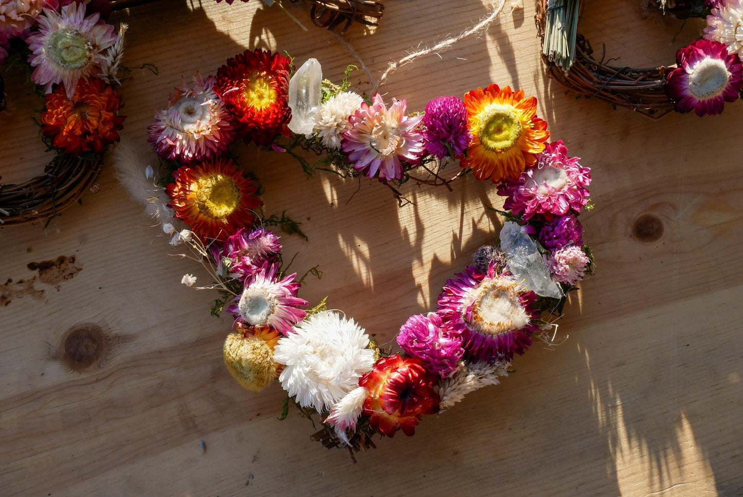 A heart-shaped wreath made of various colorful flowers, including red, orange, yellow, and white, is displayed on a wooden surface.