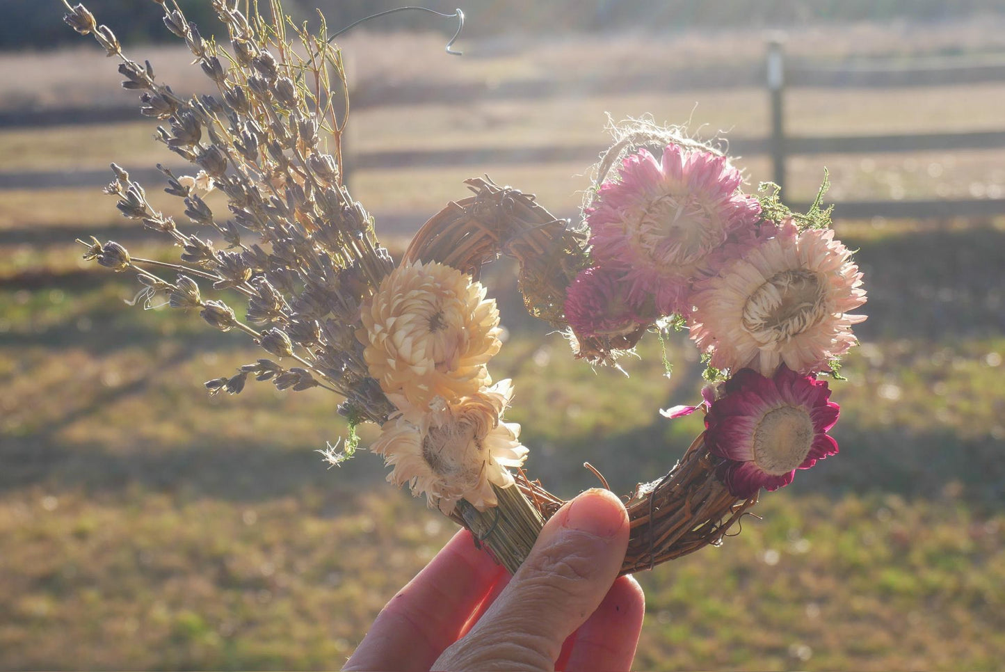 A hand holds a wreath made of dried flowers and twigs, with a field and a fence visible in the background.