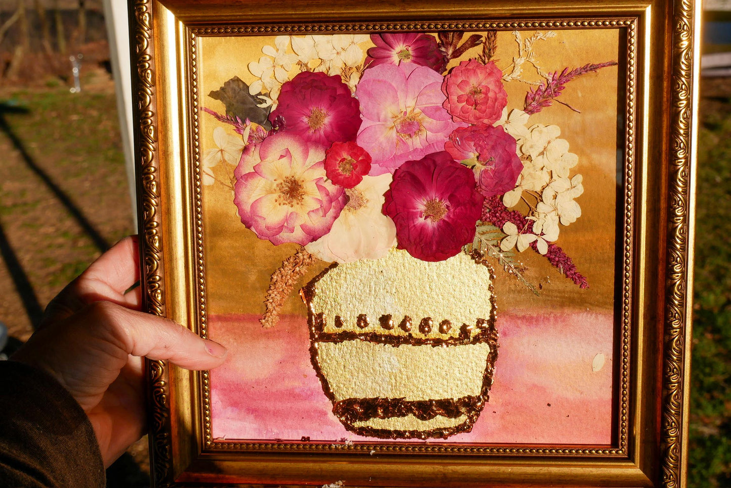 A hand is holding a framed painting of a vase filled with colorful flowers, including pink and white flowers, against a background of a garden.