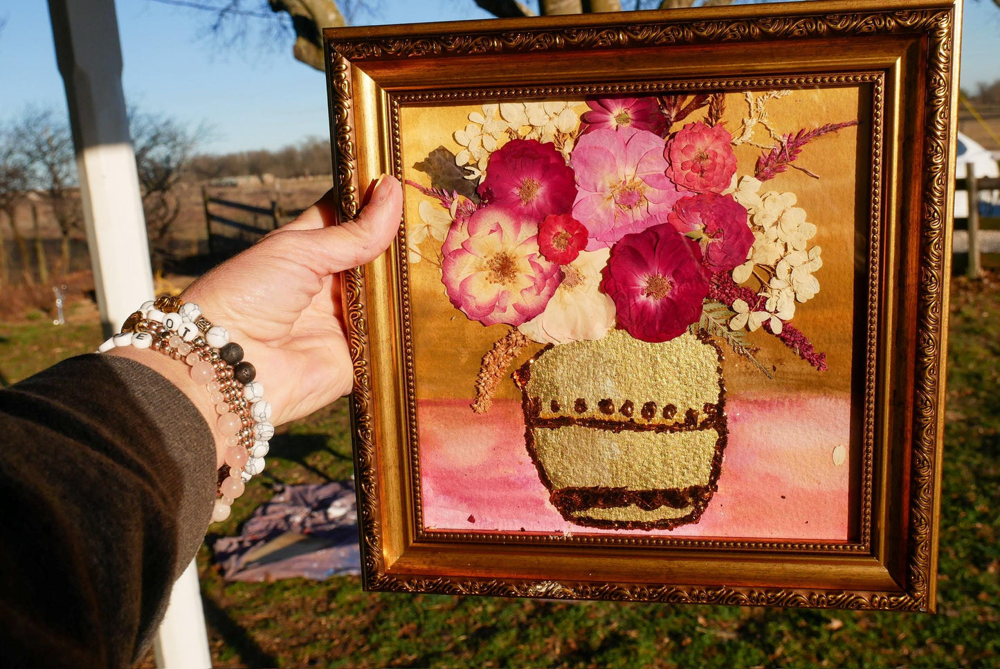 A hand holding a framed painting of a vase filled with colorful flowers, with a rural background visible.