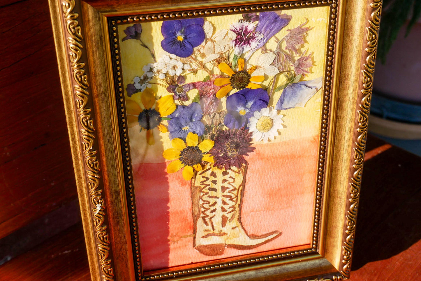 A framed painting depicting a vase filled with a variety of colorful flowers, including purple, yellow, and white blooms, set against a warm orange background.
