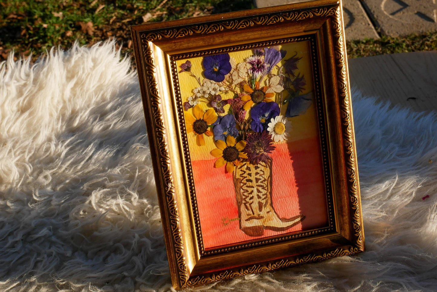 A framed painting of a vase filled with colorful flowers is displayed on a white furry surface.