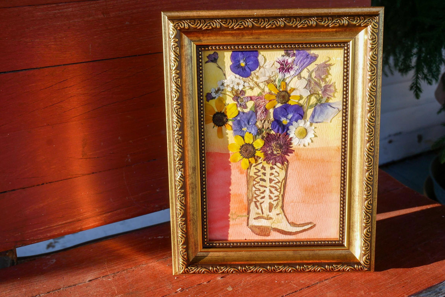 A framed painting of a vase filled with colorful flowers, including purple, yellow, and white blooms, is displayed on a wooden surface.