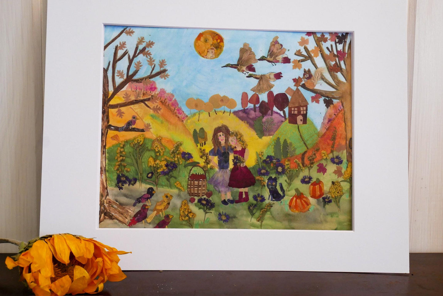 The image is a vibrant painting depicting a lively autumn scene with two girls, birds, and pumpkins. The painting is framed and placed on a surface, with a bouquet of orange flowers in the foreground.