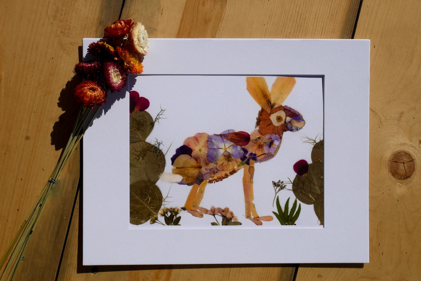 a framed artwork depicting a rabbit made from dried flowers and leaves, placed on a wooden surface.