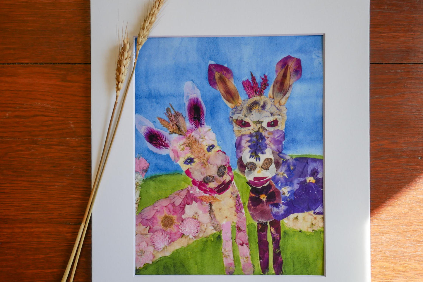 two animals, a donkey and a horse, standing together in a field with flowers. The donkey is pink and white, while the horse is purple and white. The background of the image is blue, and there are some flowers visible in the scene.