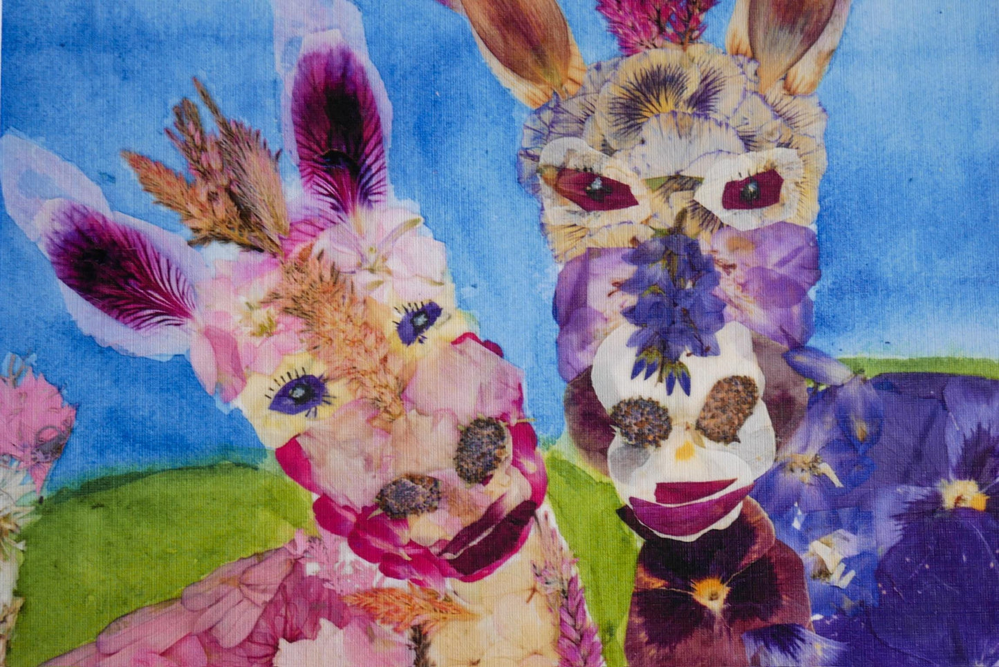 The image is a vibrant collage featuring two whimsical animal faces crafted from colorful flowers and leaves, set against a lush green field.