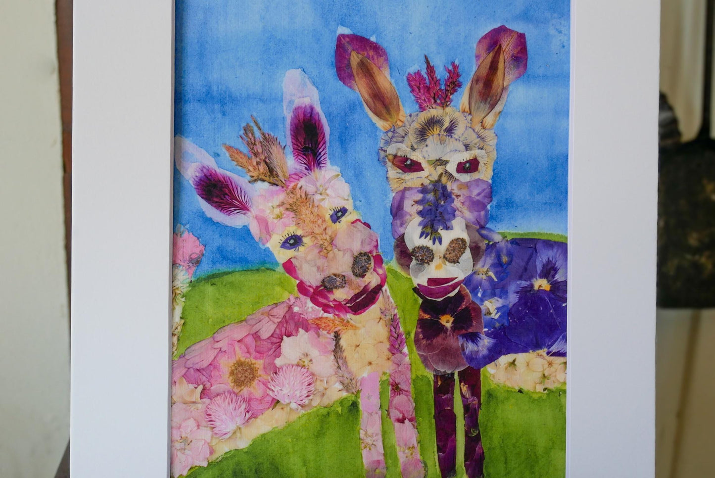 The image is a collage featuring two animals, a donkey and a horse, with their faces made of flowers. The donkey has pink and purple flowers, while the horse has purple and blue flowers. The animals are standing on a grassy field against a blue background.