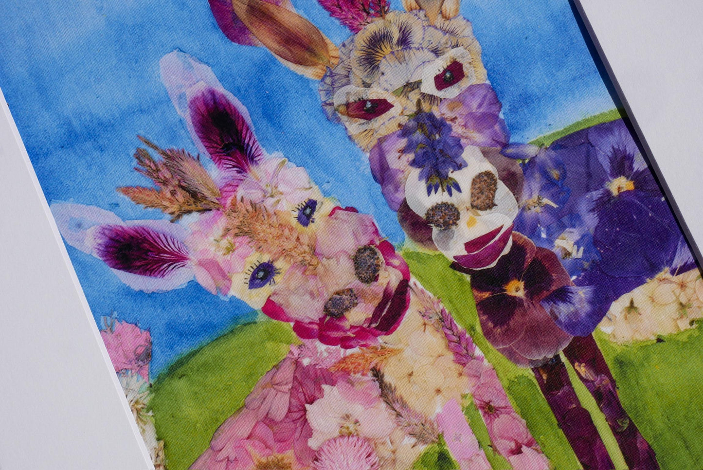 The image is a vibrant collage featuring two donkey heads adorned with colorful flowers, set against a backdrop of a lush green field and a clear blue sky.