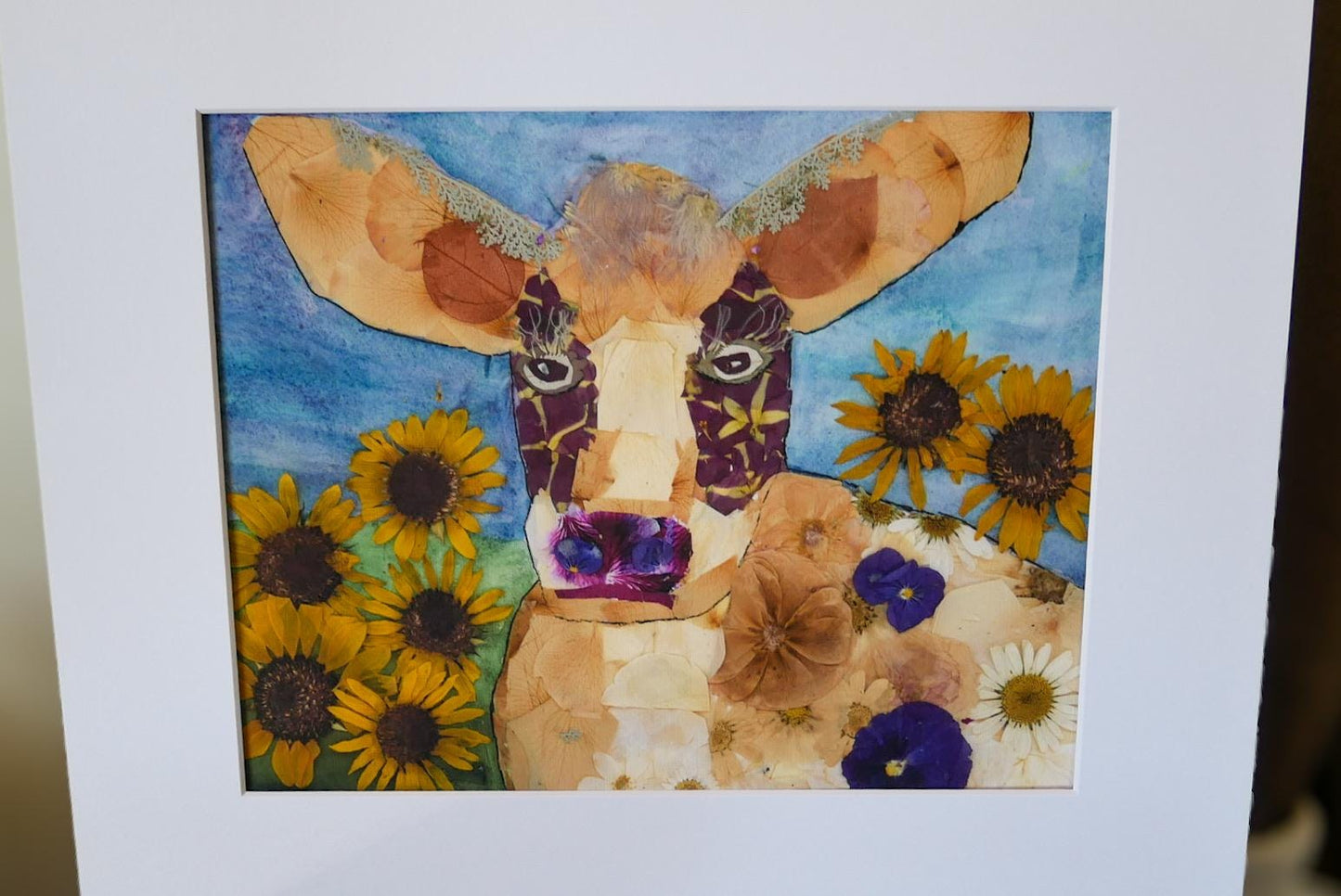 The image is a collage featuring a cow surrounded by sunflowers and other flowers, creating a vibrant and colorful composition.