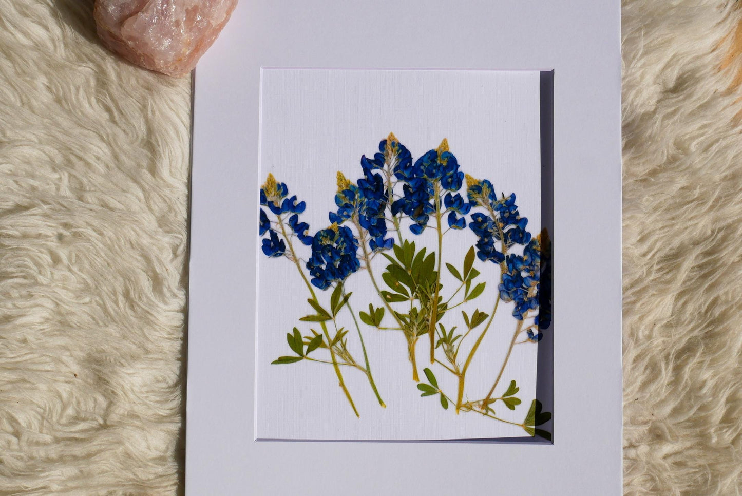 A framed image of blue flowers is placed on a white surface, with a pink crystal rock visible in the top left corner.