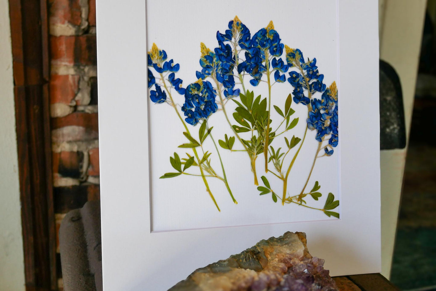 a framed artwork of blue flowers with green leaves, displayed on a table next to a rock formation.