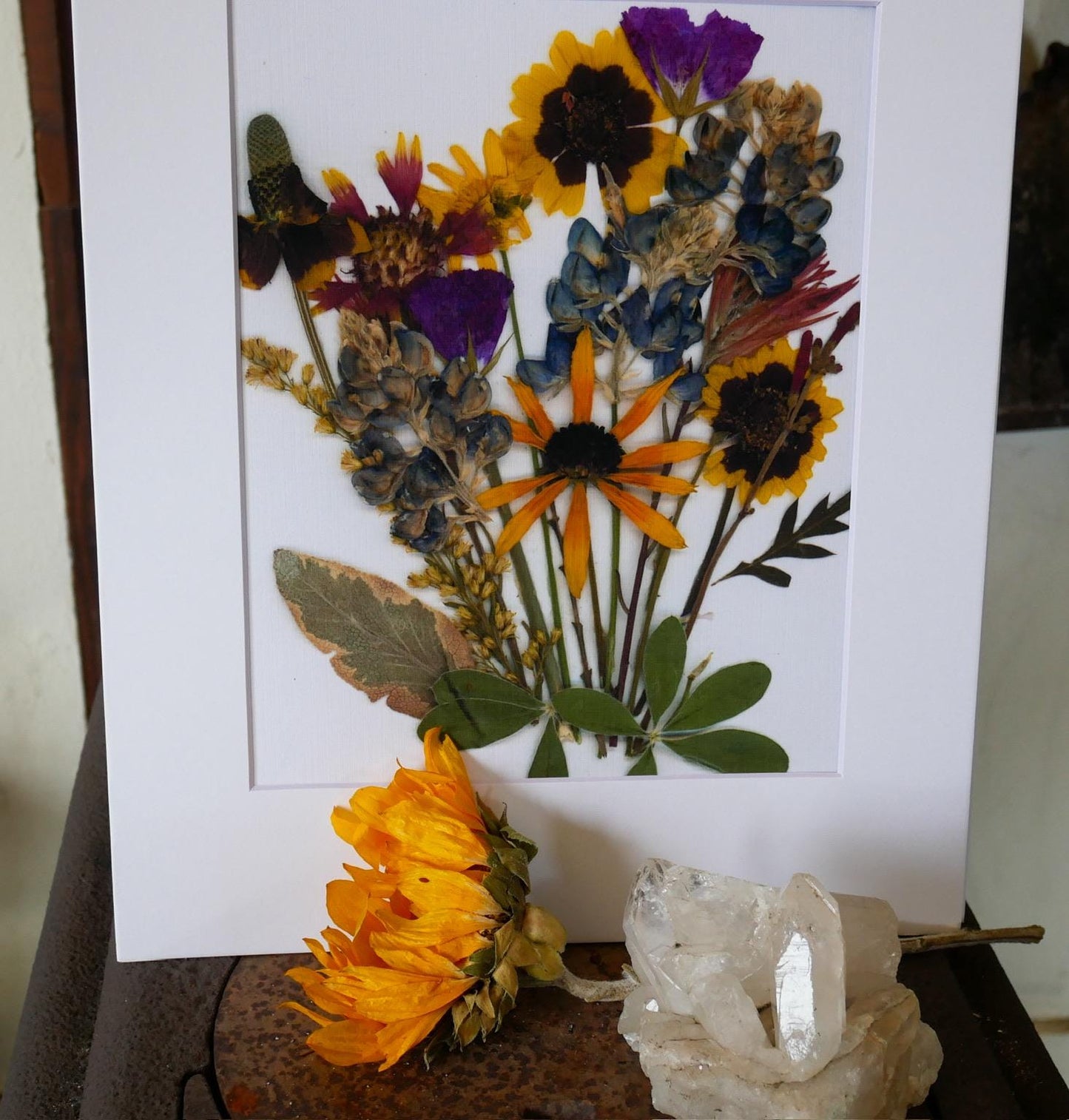 a vibrant bouquet of flowers, including sunflowers, daisies, and other colorful blooms, arranged in a white frame. The flowers are displayed on a rustic wooden surface, with a crystal formation and a single yellow flower adding to the natural aesthetic.