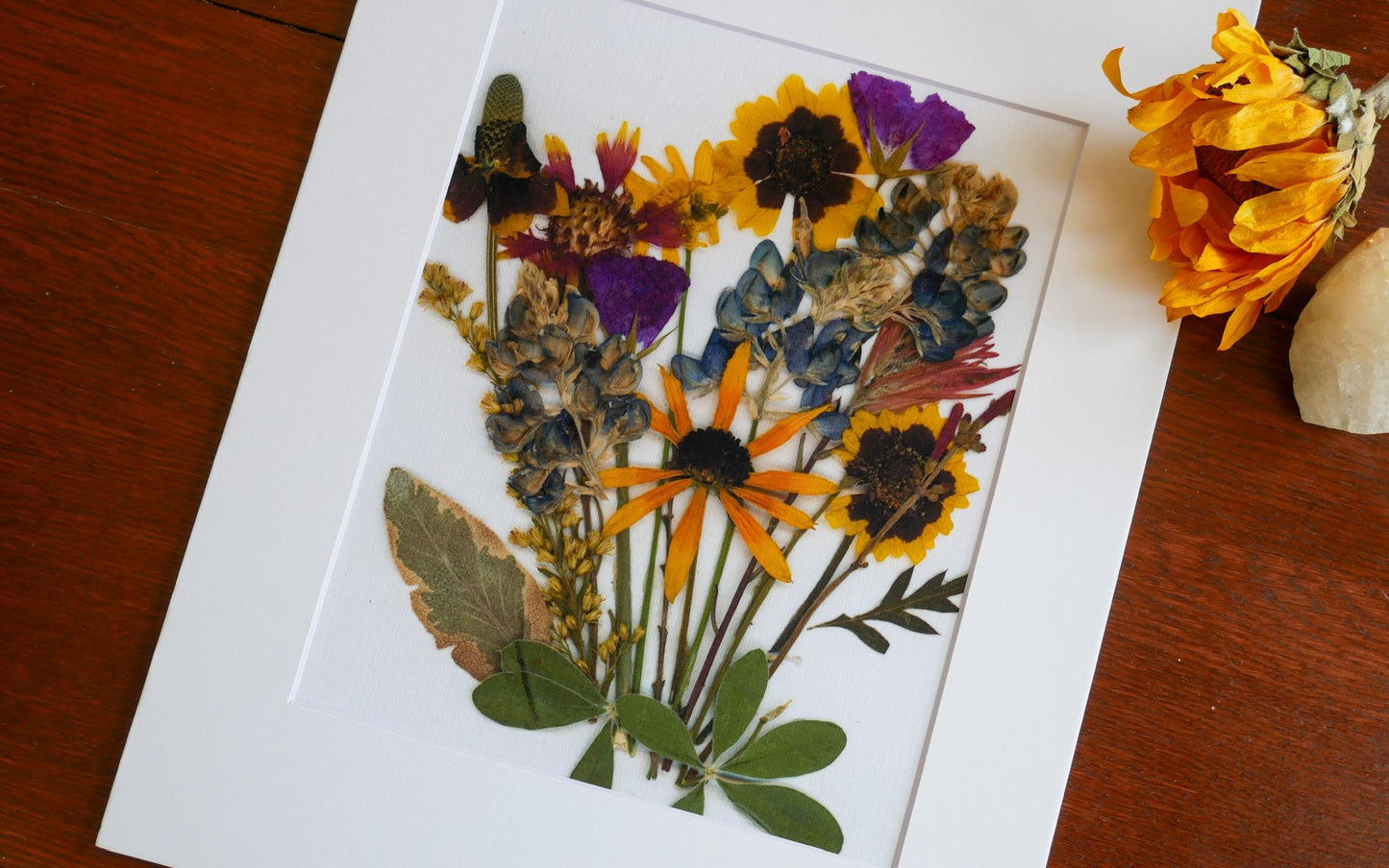 A framed arrangement of dried flowers and leaves, including sunflowers, daisies, and other colorful blooms, is displayed on a wooden surface.