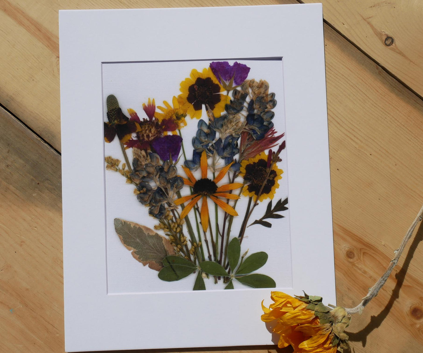 a white frame containing a vibrant bouquet of dried flowers, including sunflowers, daisies, and other colorful blooms.