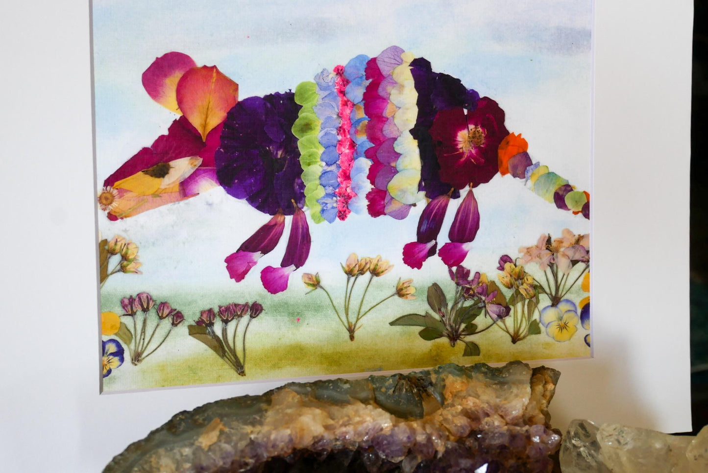 The image is a collage featuring a colorful, flower-like creature made from dried petals, surrounded by various flowers and plants.