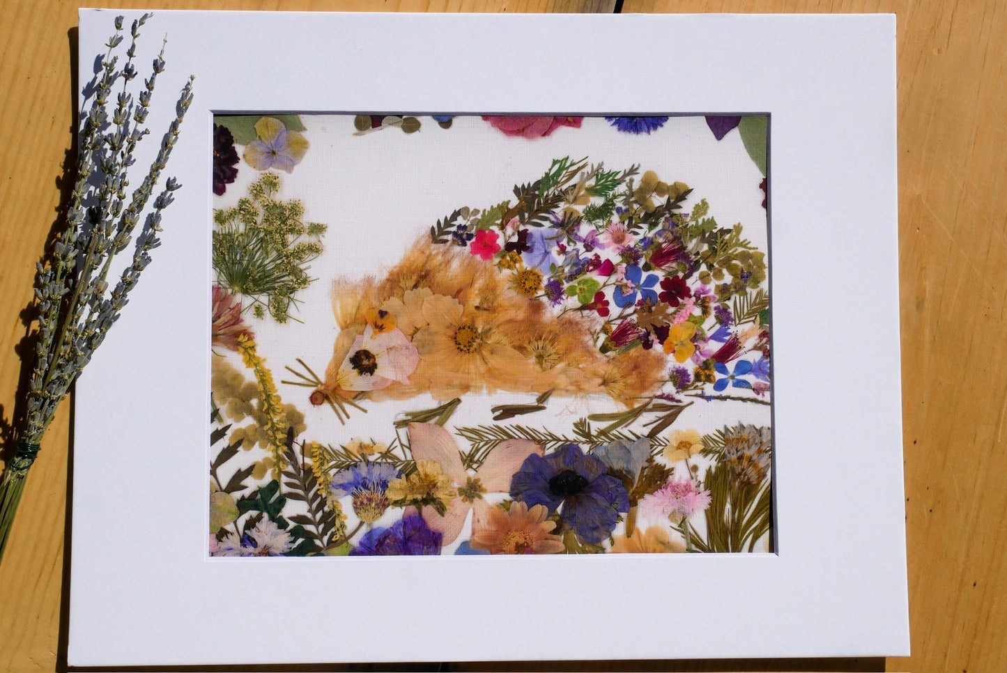 a vibrant and colorful arrangement of flowers and plants, including a variety of blooms in shades of orange, pink, purple, and yellow, as well as green leaves and stems. The flowers are meticulously arranged to create the appearance of a hedgehog, with its quills and body forming the main elements of the image. The arrangement is displayed in a white frame, which contrasts with the colorful flowers and enhances their visual appeal.