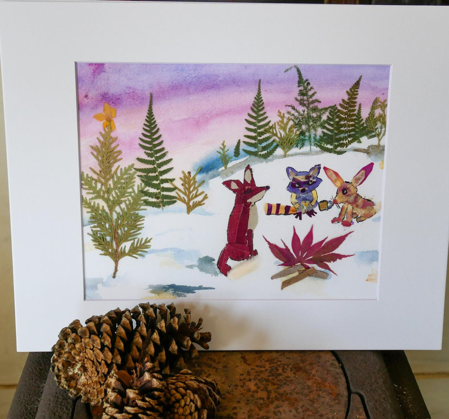 The image is a watercolor painting depicting a snowy forest scene with various animals, including a fox, a rabbit, and a deer. The painting is framed and placed on a wooden surface, with a pine cone nearby.