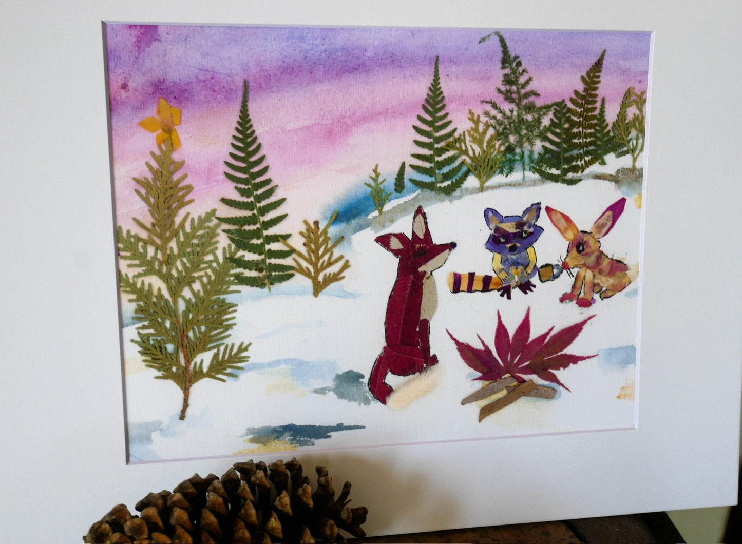 A watercolor painting depicting a snowy forest scene with three animals - a fox, a rabbit, and a raccoon - sitting around a campfire.