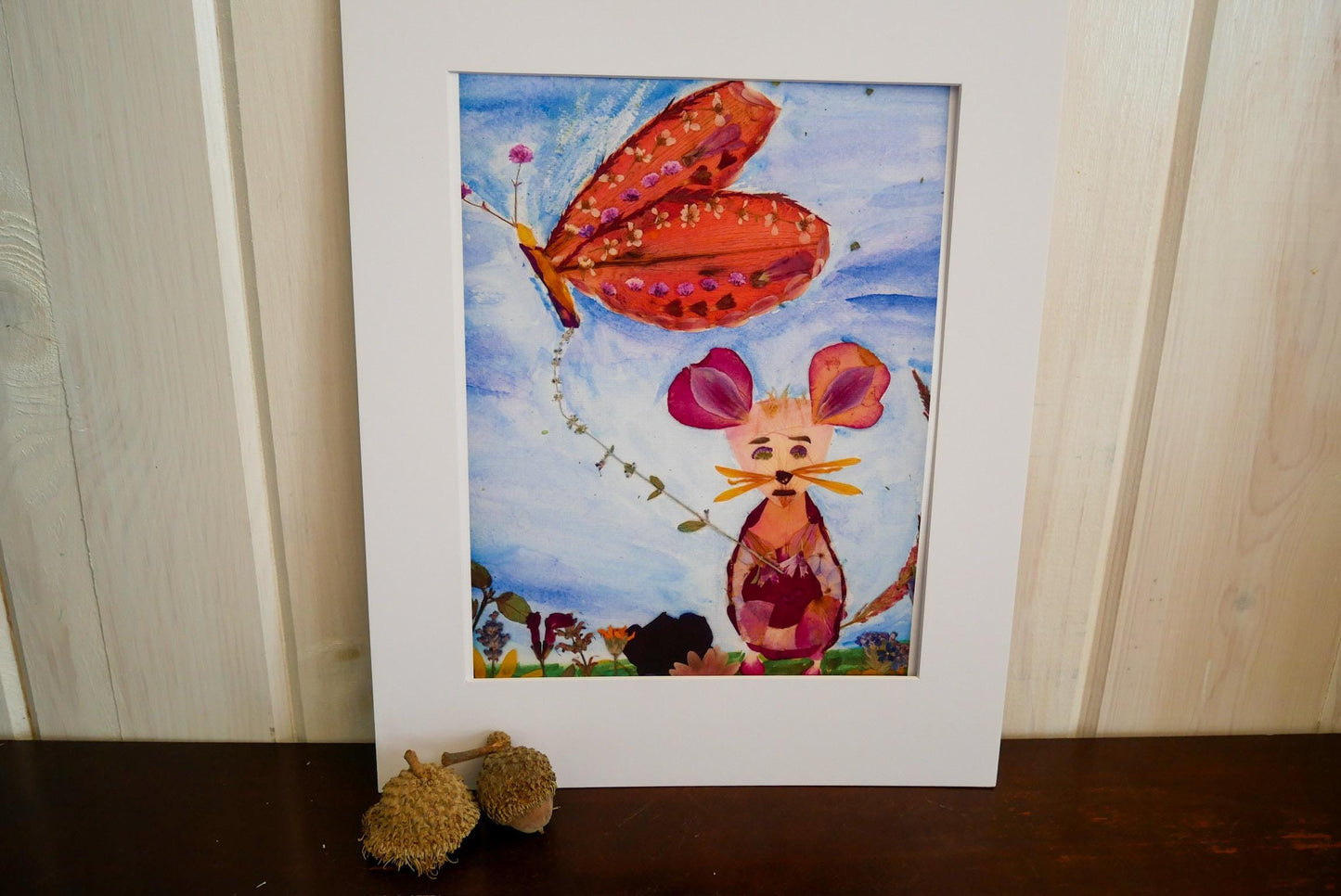 a colorful painting of a butterfly and a mouse, with the mouse holding a flower in its mouth. The painting is displayed in a white frame on a wooden surface.