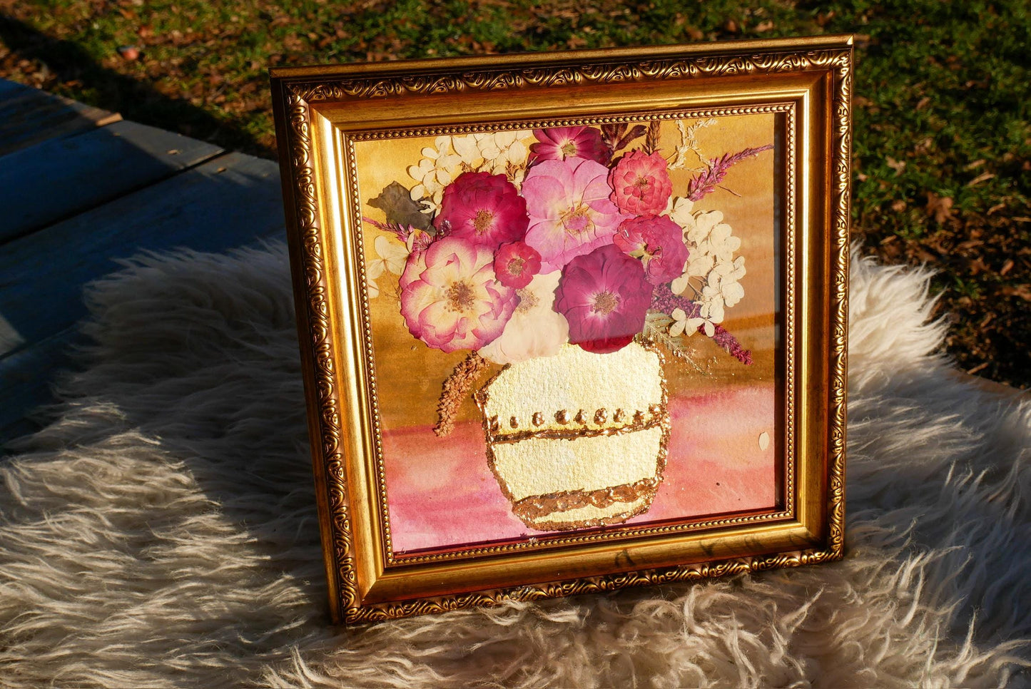 A framed painting of a vase filled with colorful flowers, including pink and white flowers, is displayed on a furry surface.