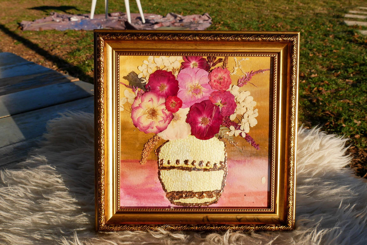 A framed painting of a vase filled with pink and white flowers is displayed on a white fur rug.