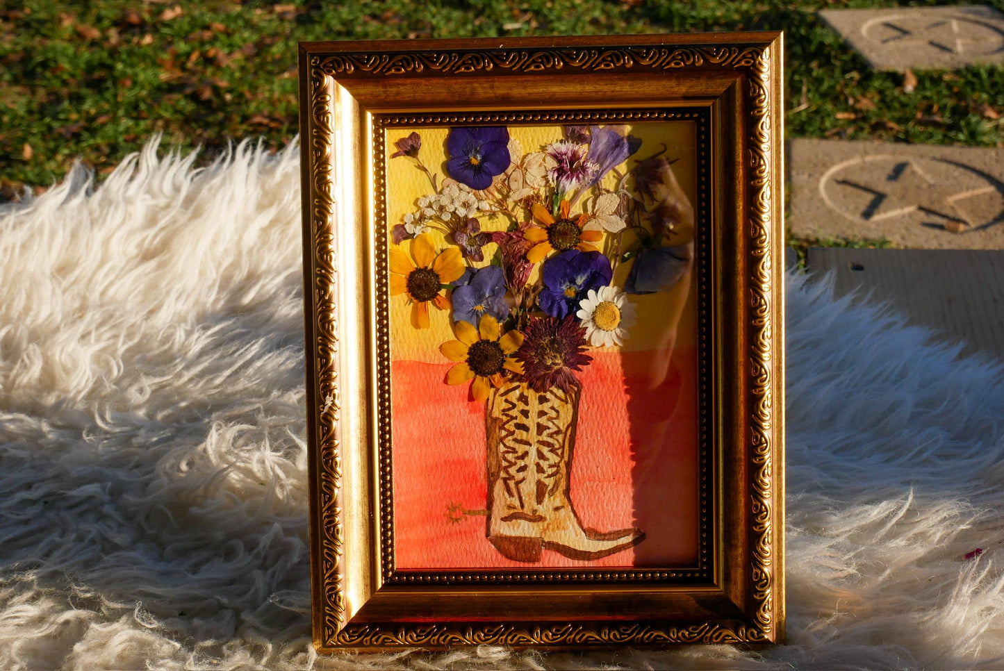 A framed picture of a vase filled with colorful flowers, including sunflowers, daisies, and other blooms, placed on a white fur rug.