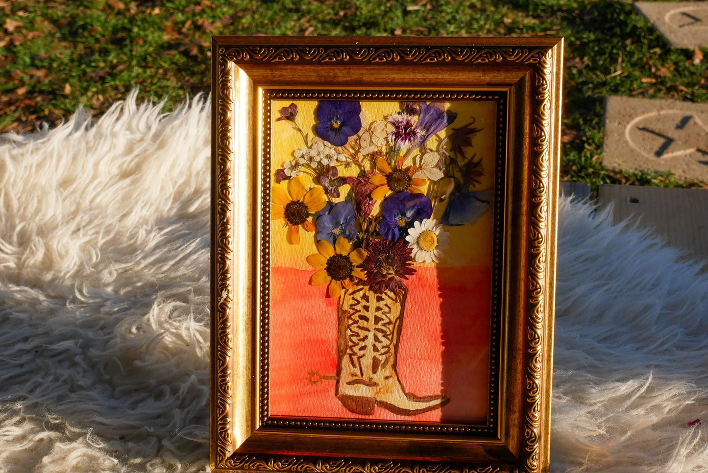 A framed painting of a flower arrangement in a brown boot, placed on a white fur rug.
