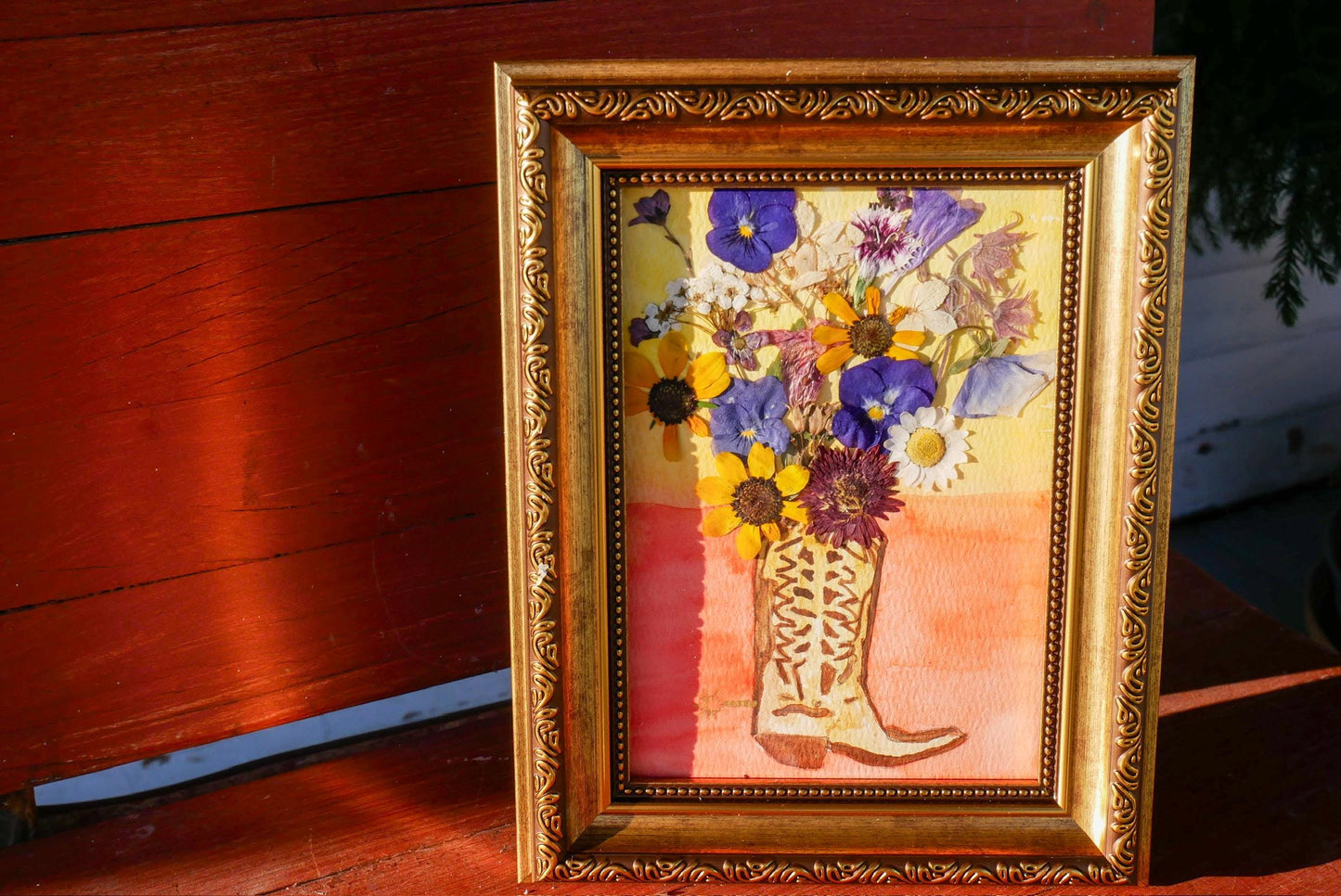 A framed painting of a vase filled with colorful flowers, including purple, yellow, and white blooms, is displayed on a wooden surface.